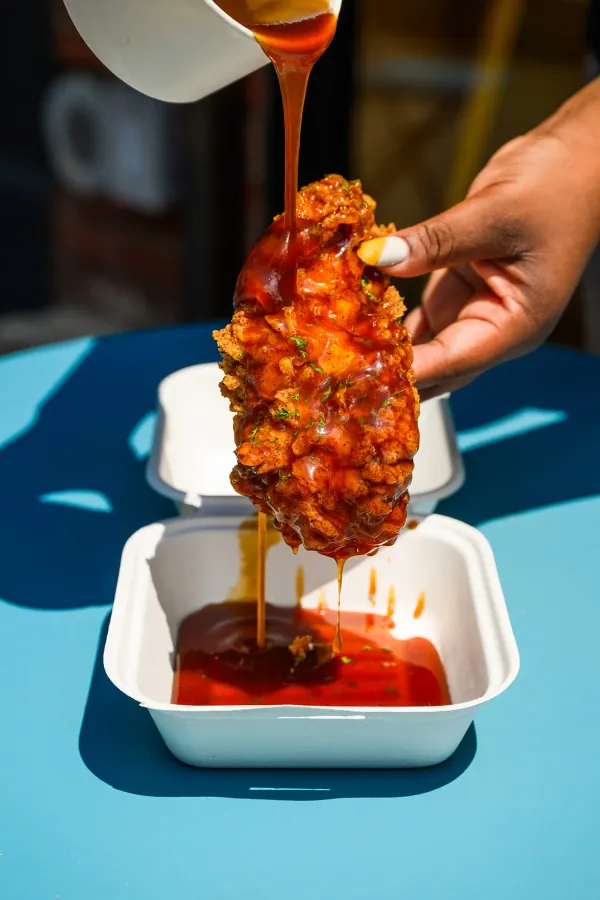 Sauce poured over a piece of fried chicken held above a takeout box on a blue table