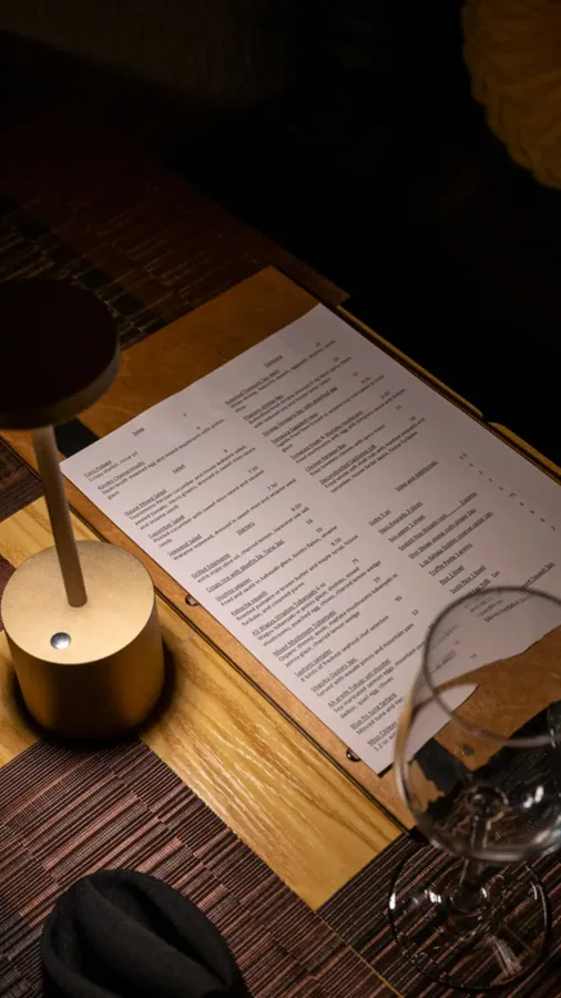 A printed menu on a wooden table beside a small brass lamp and a wine glass