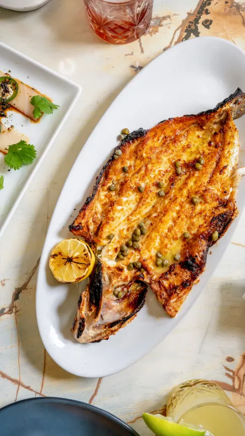 A grilled butterflied fish with capers and a charred lemon on an oval platter on a marble table