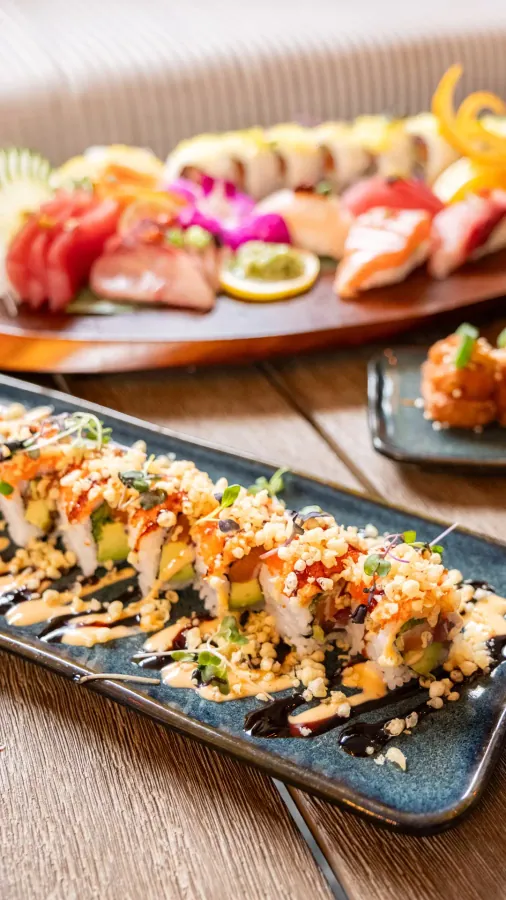 A long sushi roll topped with crispy crumbs and sauce, a sushi platter behind it on a wooden table