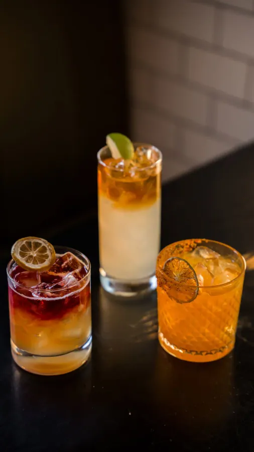 Three cocktails on a dark bar: a layered red and amber drink with a dried lemon wheel, a tall drink with a lime wedge and an orange drink in a cut-glass tumbler