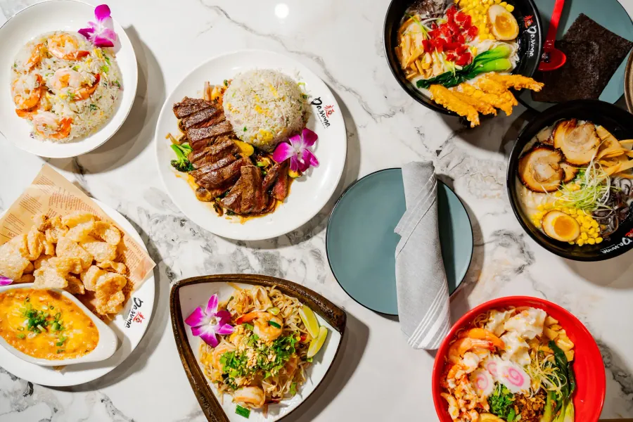 Overhead view of ramen bowls, pad thai, steak with fried rice, shrimp fried rice and a crab dip on a marble table