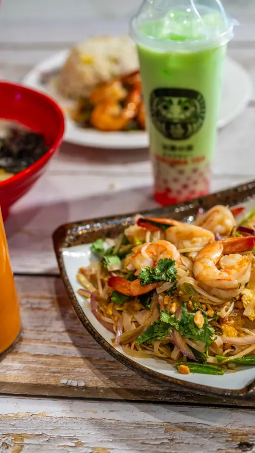 Shrimp pad thai in a triangular dish with a green bubble tea and fried rice behind it