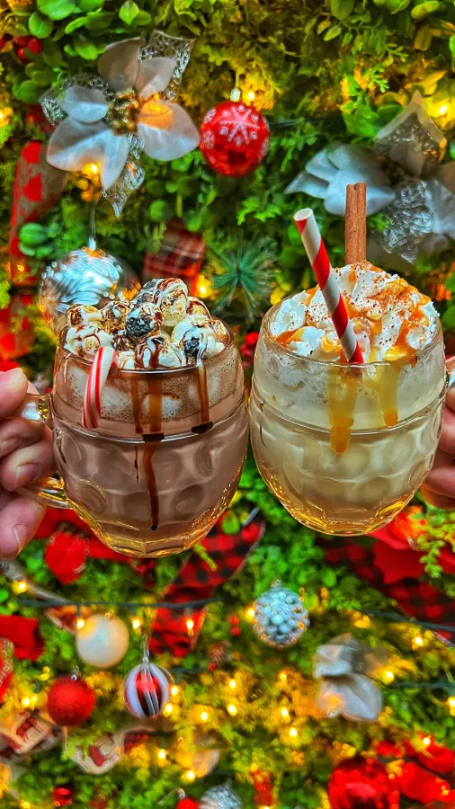 Two hands holding up hot chocolates topped with whipped cream and candy in front of a Christmas tree