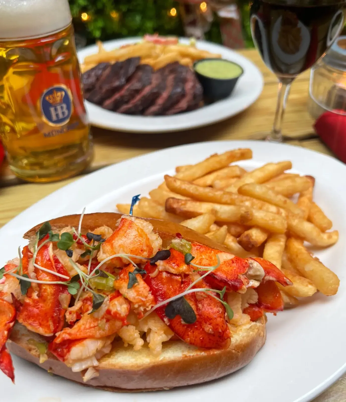 Lobster roll with fries in front of a steak plate, a beer stein and a glass of red wine